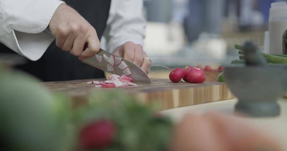 A chef chops up radishes on a cutting board outside with a city background behind him
