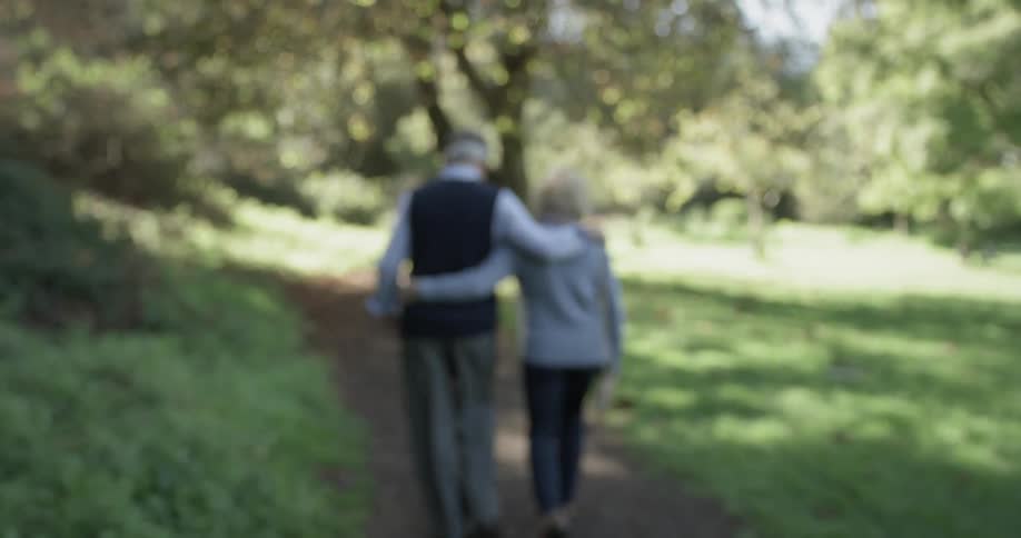 An elderly couple are walking cuddled together in the park of a sunny day