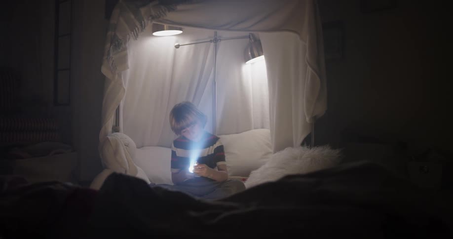 A boy messes with the beam of his flashlight inside a fort he built with lamps and sheets