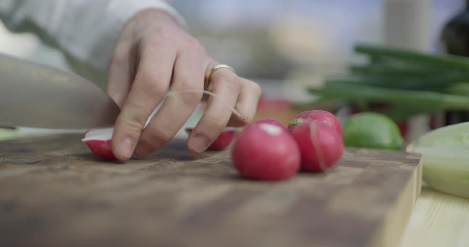 Radishes are cut on a cutting board outside on a sunny day by a chef wearing a black apron