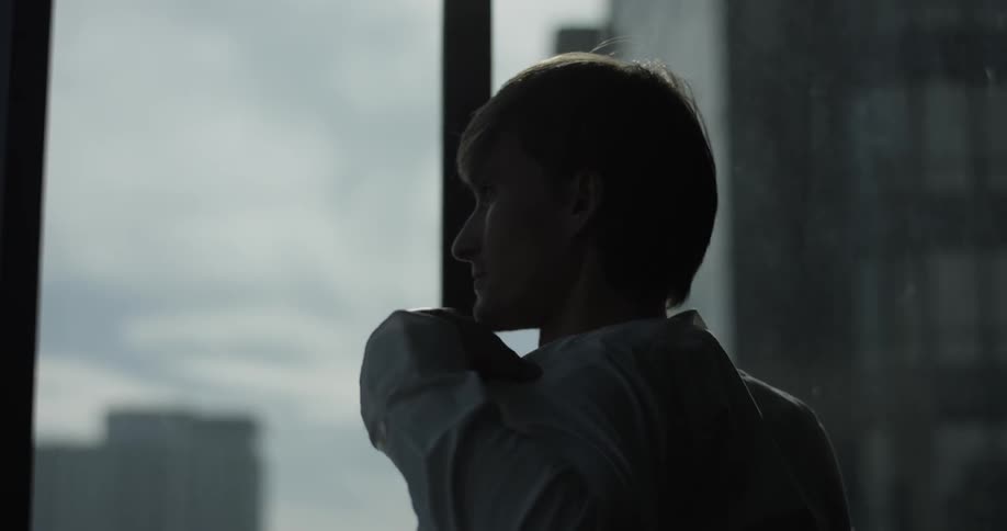 A shirtless man approached the corner of the room with giant windows overlooking the city and throws on a shirt
