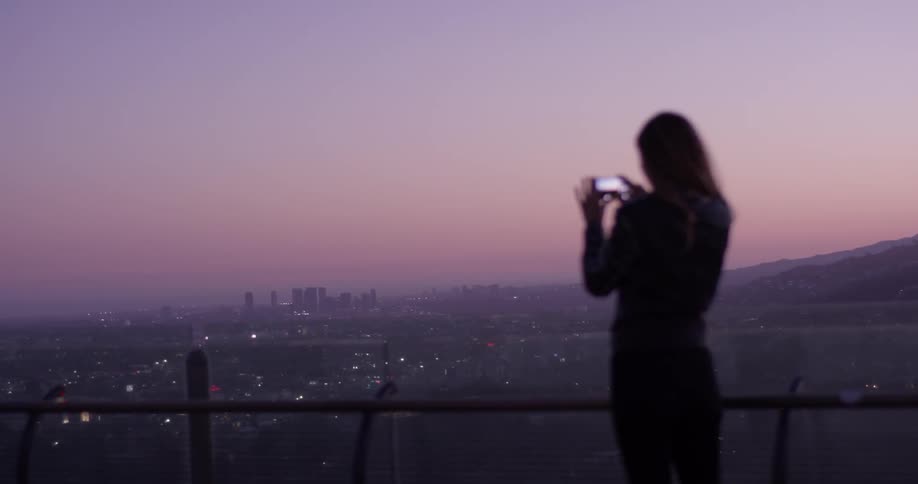 A woman takes a picture of a city with her phone as the sun sets creating a purple horizon