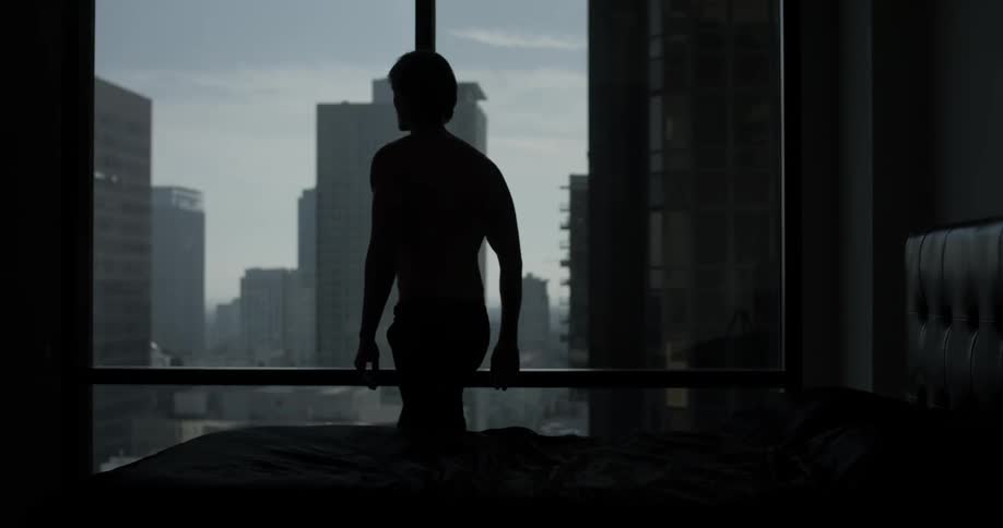 A man sits in his dimly lit room with natural lighting and stares out a window at the city he lives in