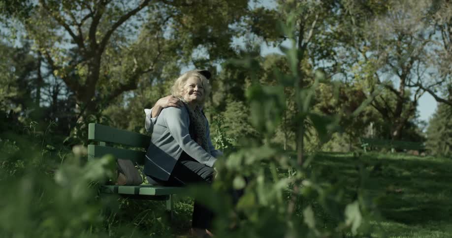 A man and a woman sit on a park bench with his arm around her on a sunny day