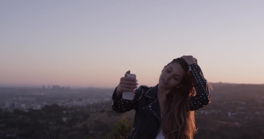 A woman is taking a selfie or a picture of herself on top of a hill as the sun sets is behind her and she adjusts her hair wearing a leather jacket
