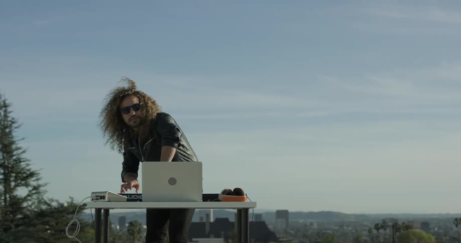 Man with long hair and leather jacket is jamming out to tunes on his laptop in the open skies of a city