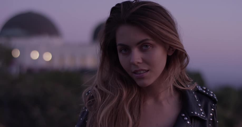 A woman blinks and looks at the camera right after the sun has set with long hair and a leather jacket with metal studs