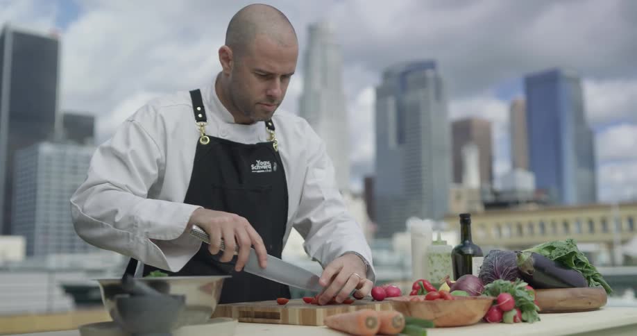 A chef stands with a skyline behind him cutting cherry tomatoes on a cutting board