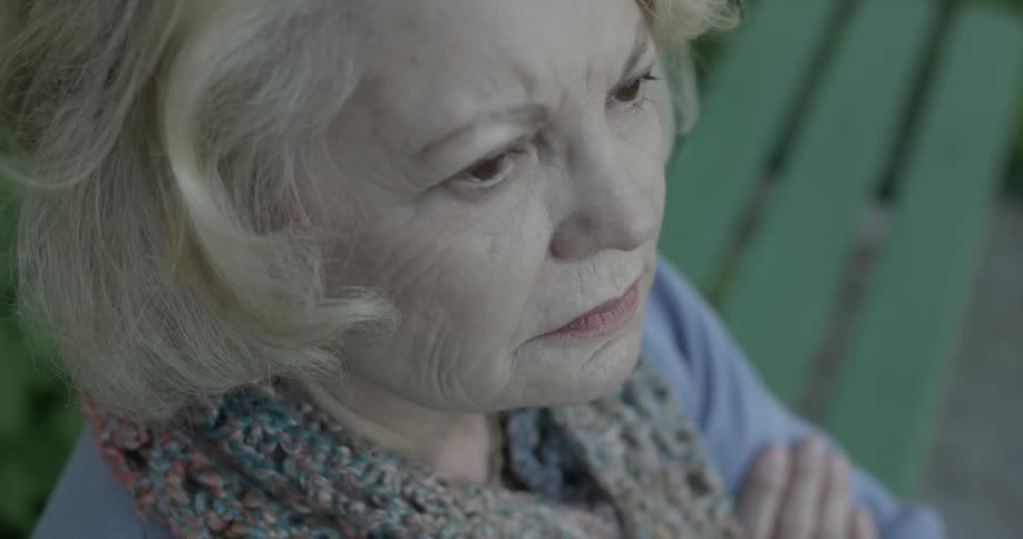 Elderly women crosses her arms while sitting on a bench on a dimly lit day looking up at the sky