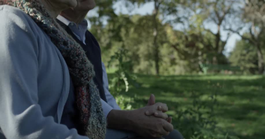 An older couple sit on a bench and clasp hands together