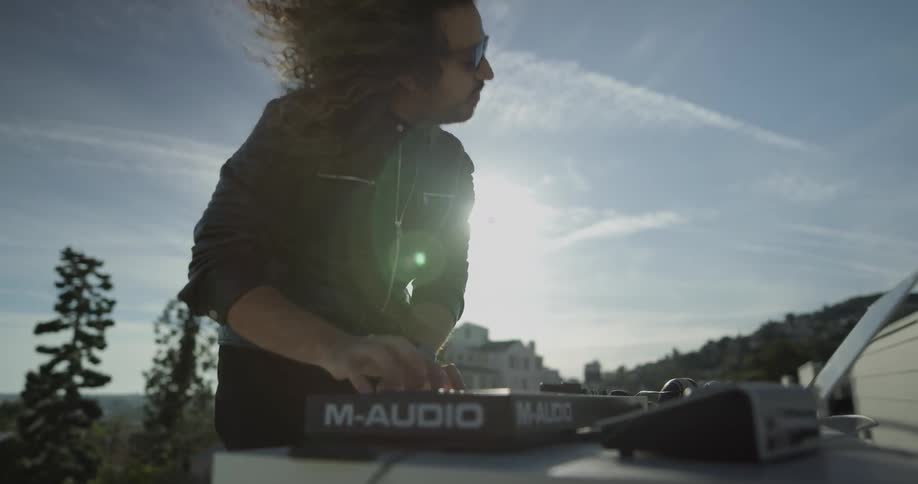 A man does a dj performance outdoors with his laptop and other djing equipment on top of a table
