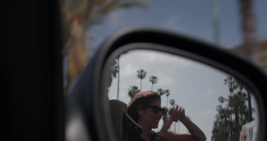 A side mirror of a convertible shows a woman dancing in the back seat