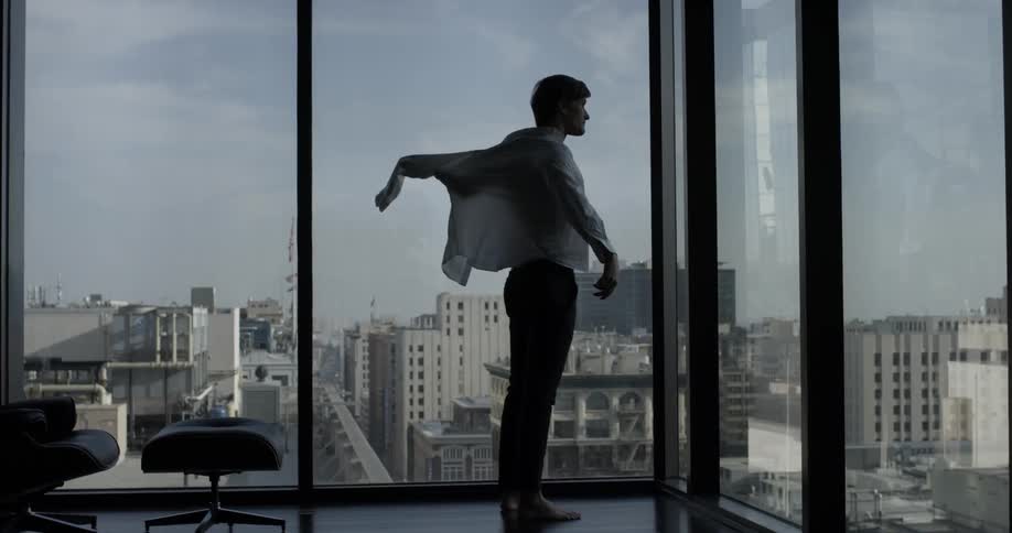 A man walks towards a window overlooking a city and puts on a shirt