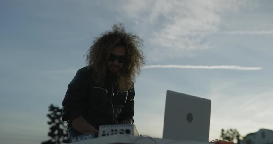 A man stands in front of a table with dj equipment outside on a sunny day near sunset