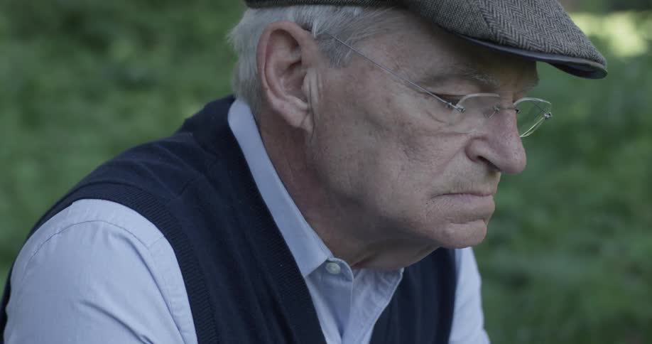 An old man in a hat and glasses stares at something off screen