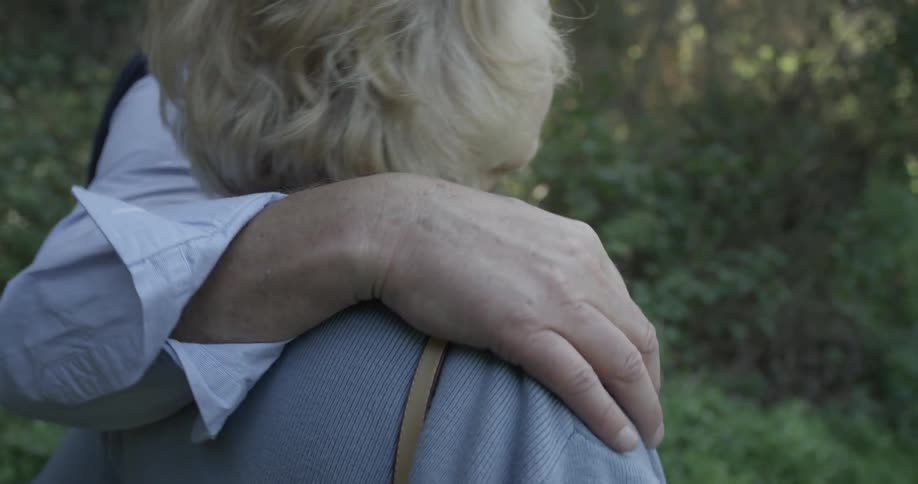 An elderly couple stroll through the woods or a park and seem to be content being with one another