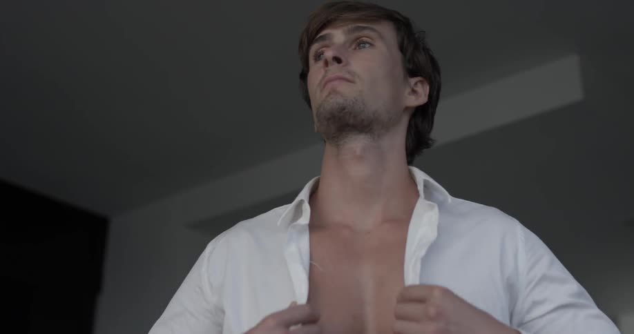 Man puts on a shirt and buttons it up as he looks out at something in the distance