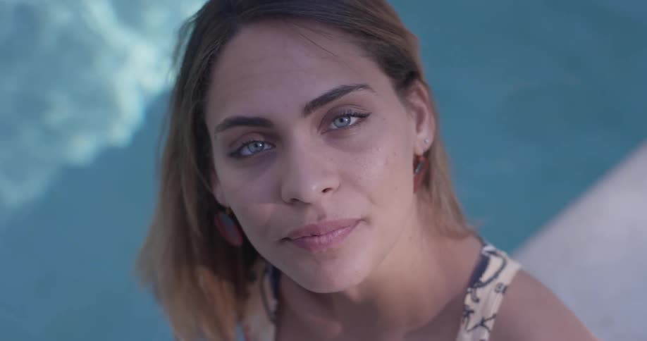 A woman sitting beside a pool, brushes her hair behind her ear