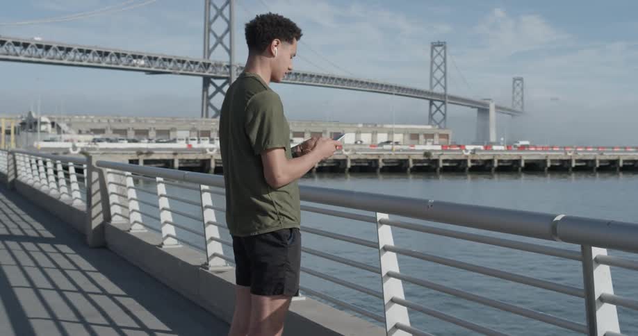 A man walks over to the railing fence gate of the ocean pier and marina and stands there pulling out his phone looking off into the distance wearing air pods