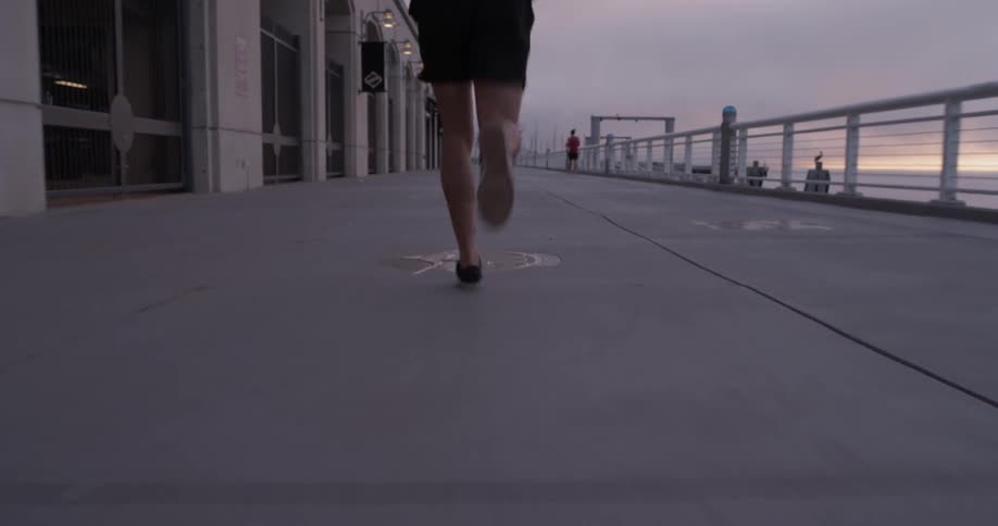 Man is jogging down the sidewalk of sunset day near the oceanfront and a bridge and buildings running on the cement