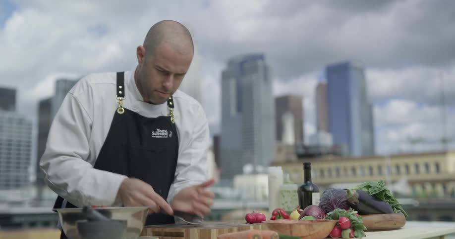 A man scoops up tomatoes with a knife and puts them in a bowl on top of a roof overlooking the city