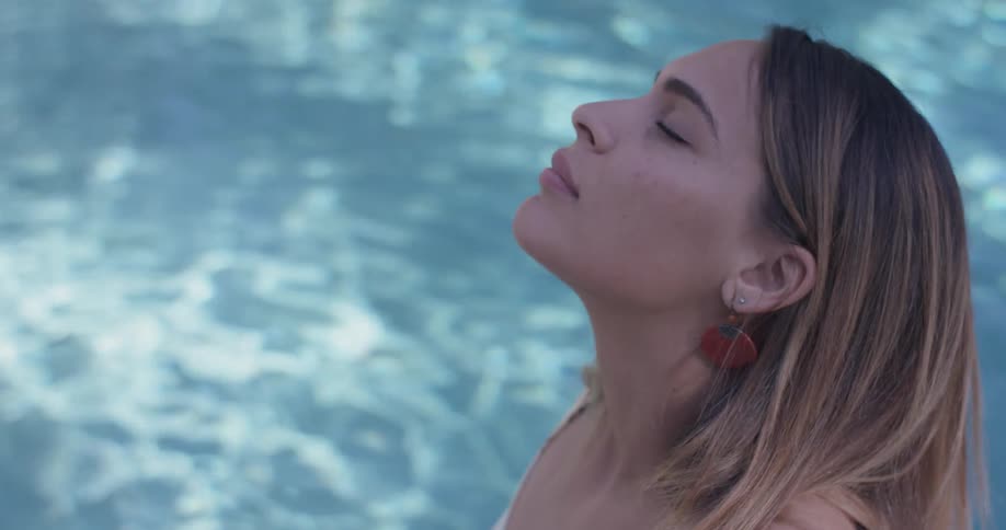 A woman in a pool tilts her head back and move side to side relaxing as she stands on a sunny day