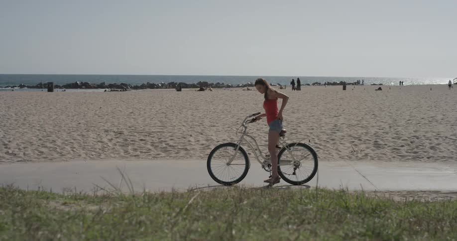 Woman rides her bike on a sidewalk by the beach and stops to take out her phone