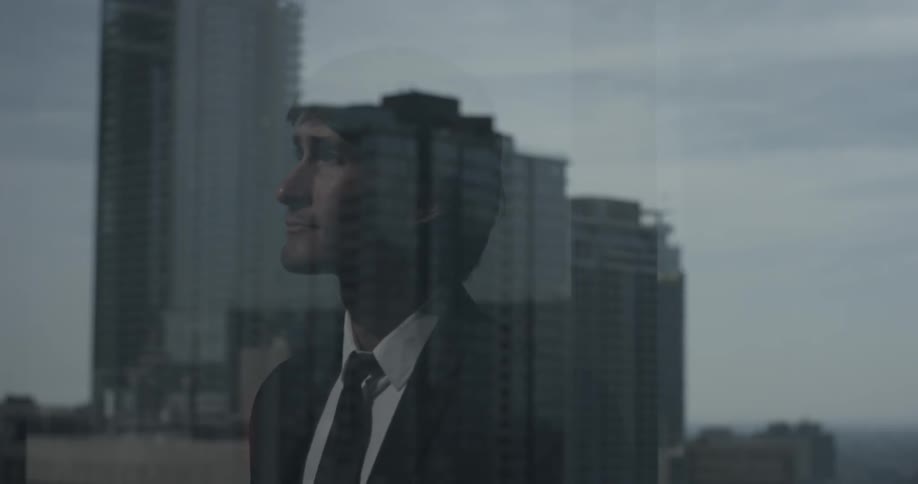 A man can be seen in the reflection of a window showing a large downtown building and cloudy skies