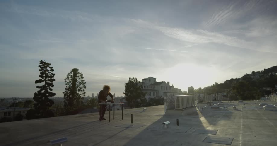 A man is outdoors in front of a table on a roof with his laptop and dj equipment