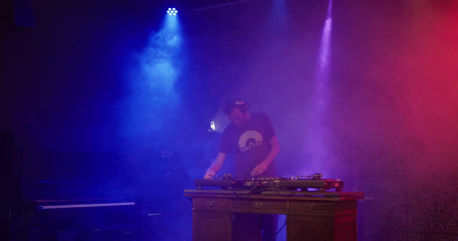 A man putting on his headphones at a dj booth with blue and red lights in the background