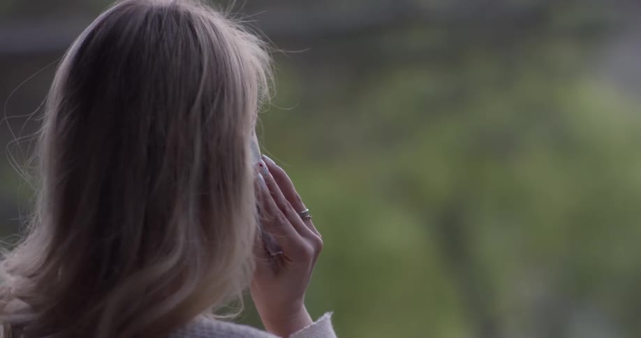 A blond woman holds a cell phone up to the ear and talks in it while outdoors in nature on a sunny day