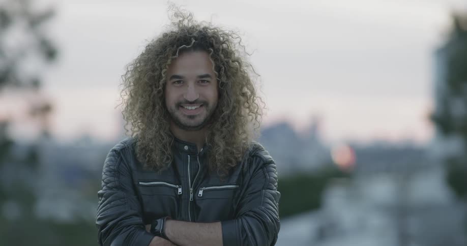 A man smiles at the camera with his long hair and leather jacket arms crossed on a scene where the sun is setting