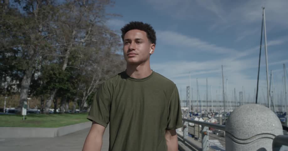 Man puts in his air pods while walking down the sidewalk at the marina on a bright sunny day