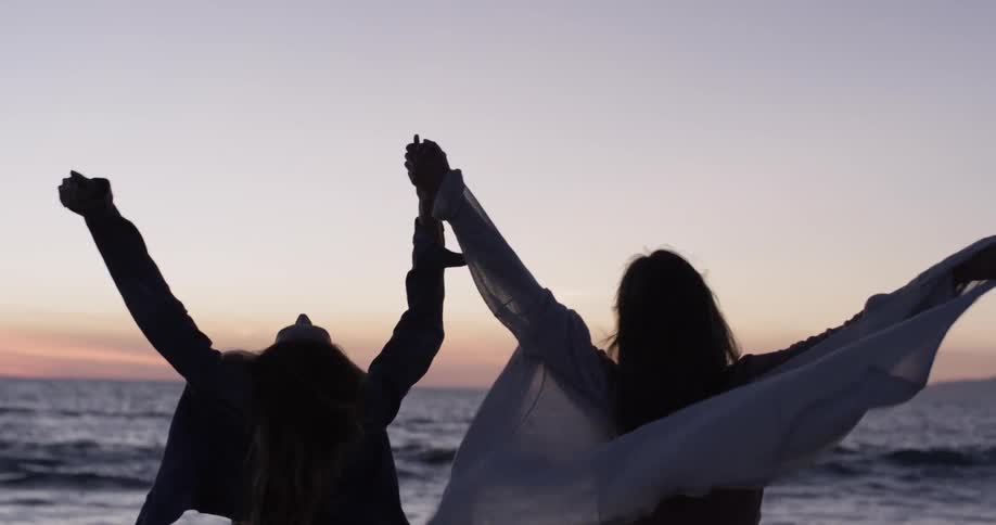 Two females holding hands on a beach throwing their hands up in the air