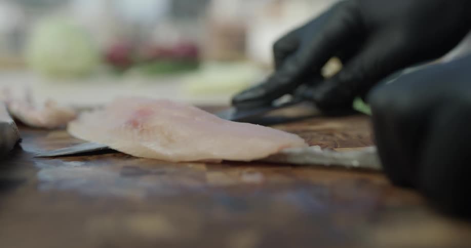 A chef fillets a cut of fish on a cutting board with a sharp knife and black gloves on a sunny day outside
