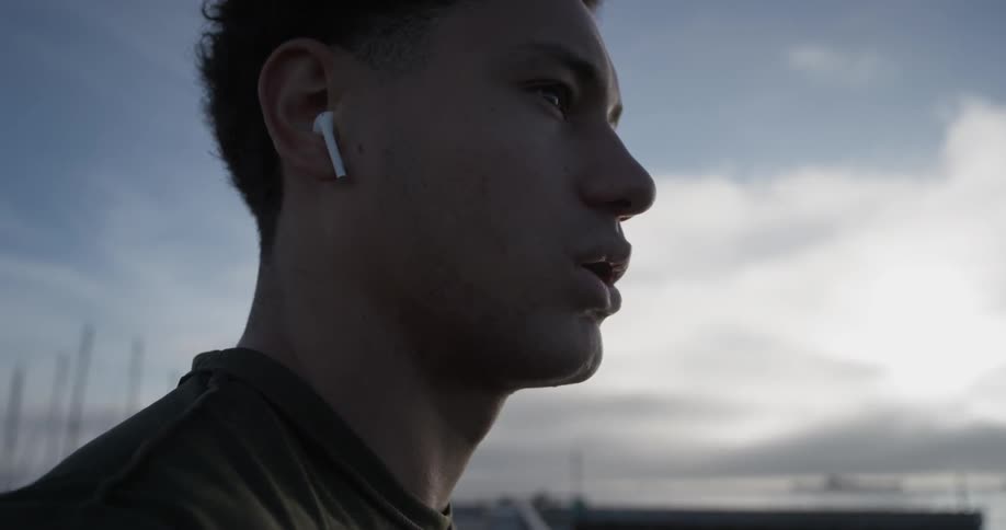 A man has earbuds in his ears and stands looking out at the horizon of a sun setting cloudy sunny day and he looks exhausted as he breathes