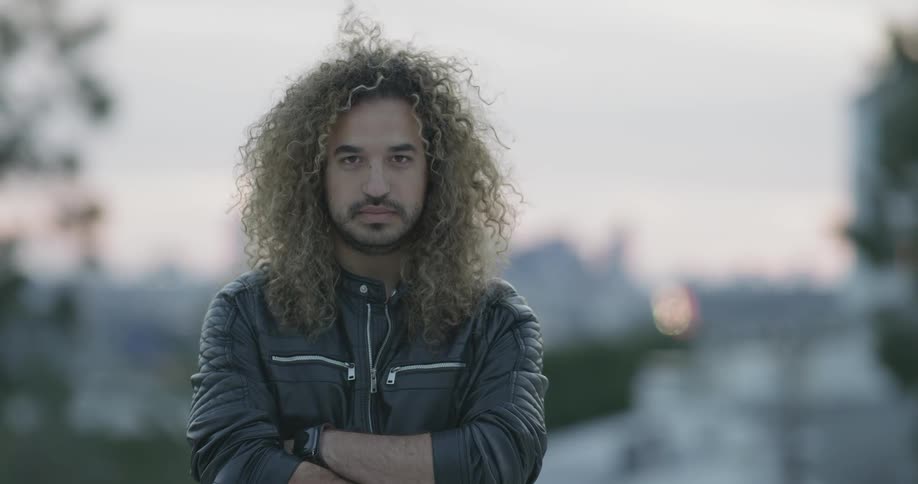 A man stands there and opens his eyes wearing a leather jacket and gnarly hair with light facial hair