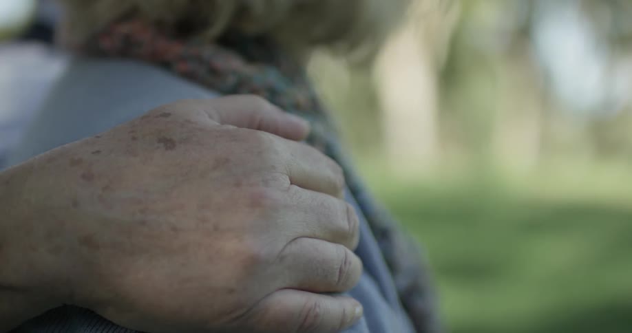 Elderly hand is on the shoulder of an old women on a park bench in the city with a bright sunny day lighting up the scene