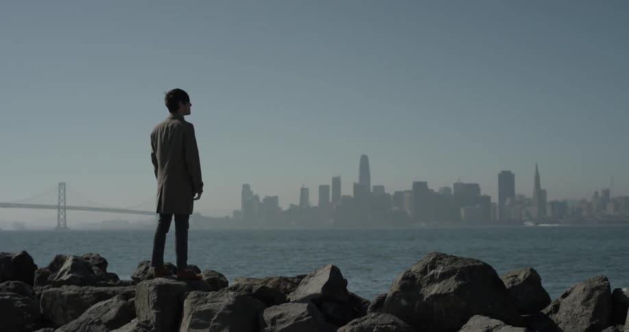 Man stands on large rocks looking out at a body of water and a city