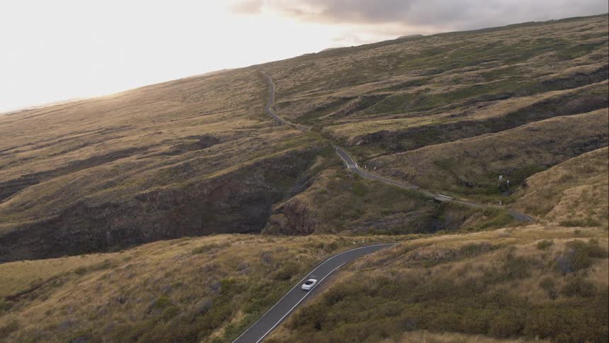 A lone car drives on a long road through the hillside at noon