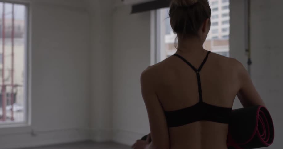 A woman walks into an empty room and unrolls a yoga mat