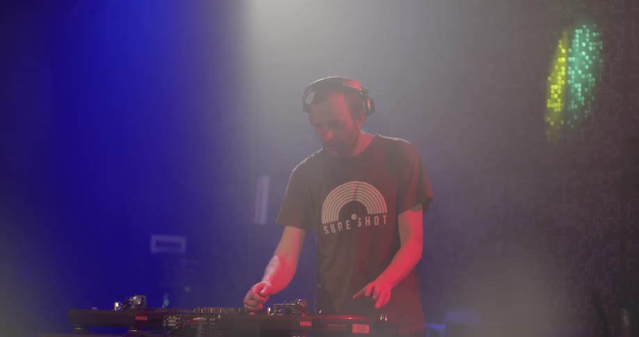 A dj wearing headphones stands in front of his equipment as lights flash across the room