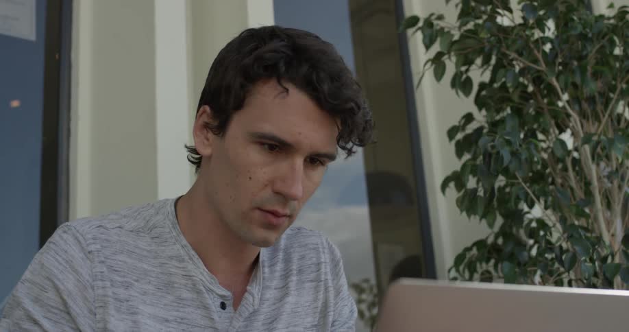 A man sits outside typing on a laptop as his hair blows in the wind