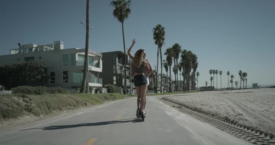 Two woman on scooter ride on sidewalk next to beach
