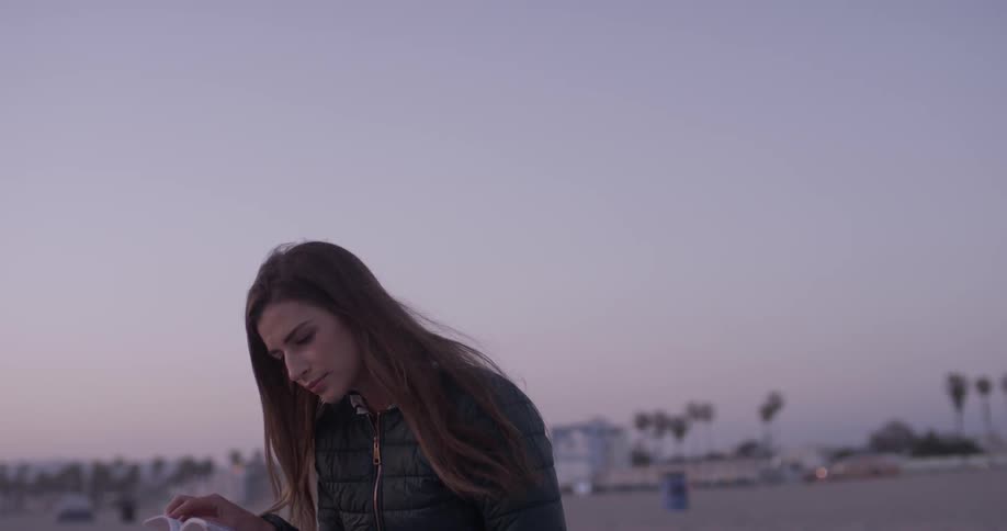 A beautiful long haired women with a puffy jacket read a book at the beach after sunset