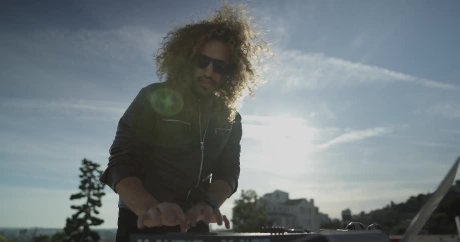 A man is outside with a table in front of him which has all his dj equipment and he is bobbing his head while doing a performance