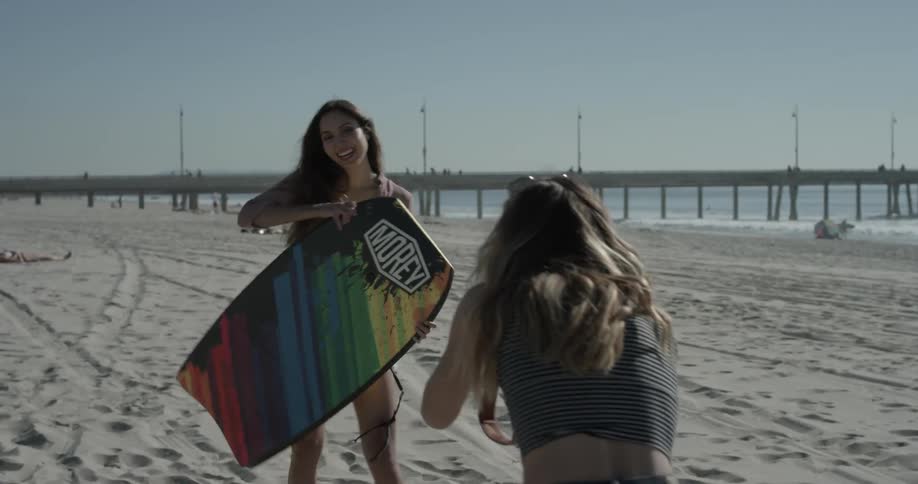 A woman takes a picture of another woman holding a surfboard on the beach