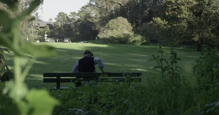 Man walks through the park and tips his hat approaching a bench and sits down placing his book down and stares out at the park