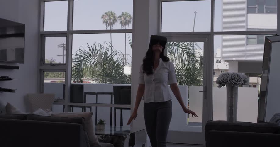 A woman is standing with virtual reality goggles on in her living room and seems to be playing a game on the device