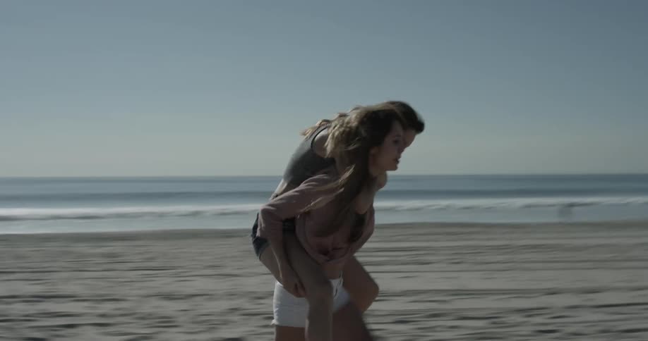 Piggy back ride between two cute youthful girls wearing shorts and excitedly walking along the sand of the beach